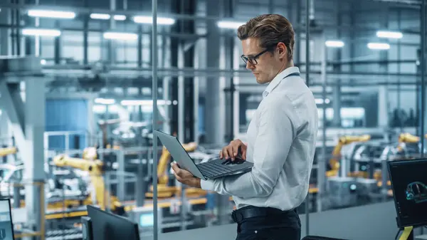 在一家汽车组装厂 一位戴眼镜和白衬衫的英俊工程师正在使用笔记本电脑 并从办公室向外张望 技术设施车辆设计工业专家 — 图库照片
