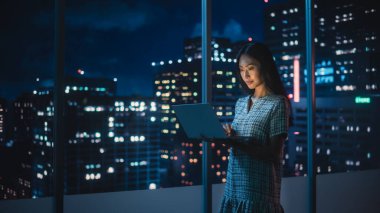 Finans Analisti Laptop Bilgisayar Kullanıyor, Gökdelenli Güzel Gece Şehri Skyline 'ı olan Pencerenin yanında duruyor. Modern Kurumsal Ofiste çalışan kendinden emin Asyalı İş kadını.