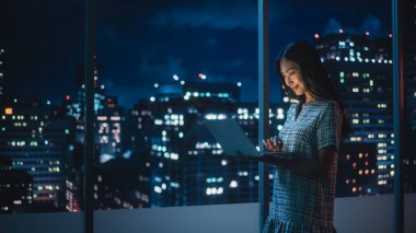 Finans Analisti Laptop Bilgisayar Kullanıyor, Gökdelenli Güzel Gece Şehri Skyline 'ı olan Pencerenin yanında duruyor. Modern Kurumsal Ofiste çalışan kendinden emin Asyalı İş kadını.