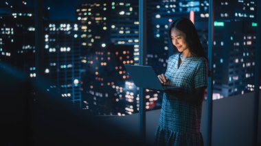 Finans Analisti Laptop Bilgisayar Kullanıyor, Gökdelenli Güzel Gece Şehri Skyline 'ı olan Pencerenin yanında duruyor. Modern Kurumsal Ofiste çalışan kendinden emin Asyalı İş kadını.