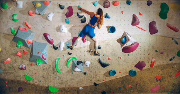 Спортивная женщина-скалолаз практикующая сольное восхождение на каменную стену в тренажерном зале. Женские упражнения в помещении фитнес-центра, занятия экстремальным спортом для ее здорового образа жизни. Выстрел со спины.