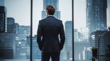 Confident Young Businessman in a Perfect Tailored Suit Standing in His Modern Office Looking out of the Window on Big City with Skyscrapers. Successful Finance Manager Planning Project Strategy. clipart