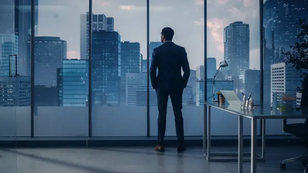 現代のオフィスに立つ完璧なテーラードスーツを着た思慮深い若いビジネスマン夕方の大都市の窓から外を見る 成功した財務マネージャー計画プロジェクト戦略 — ストック写真