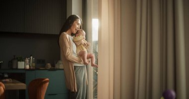 Güzel, Asyalı bir kadın, bebeğini kollarında tutarken evinde bir pencerenin yanında duruyor. Sevimli Küçük Bebek Annesinin Kucağında Dinleniyor