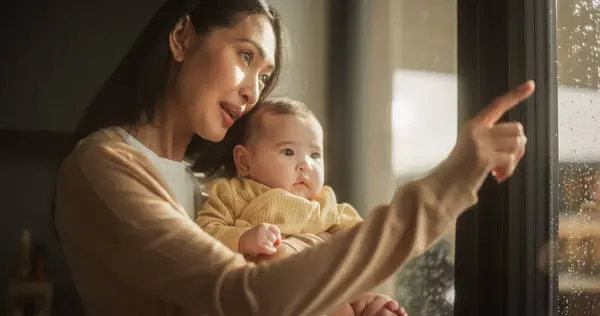 美しい若いアジアの女性は 自宅の窓の隣に立っている間 彼女の腕に赤ん坊を抱いています かわいい リトル トッドラー 彼女の — ストック写真