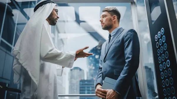 Liikemies Puhuu Arabialaisen Sijoituskumppanin Kanssa Matkustaessaan Lasihissillä Toimistoon Modernissa Liikekeskuksessa — kuvapankkivalokuva