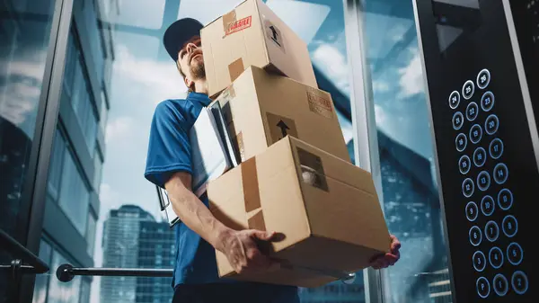 Young Delivery Person Riding Glass Elevator in Modern Office Building. Mail Courier Holding Cardboard Parcel Boxes. Handsome Mailman Delivering