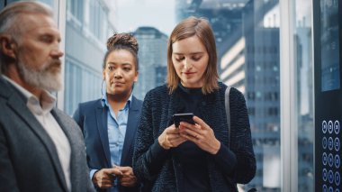 Kendine güvenen genç yetişkin kadın cam asansörle modern iş merkezindeki ofise gidiyor. Başarılı Yönetici Akıllı Telefon Kullanımı, Mesaj Yazma, Denetim