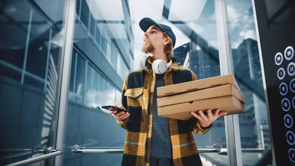Young Food Delivery Person Riding Glass Elevator in Modern Office Building. Restaurant Courier Holding Take Away Pizza Boxes. Handsome Employee Using