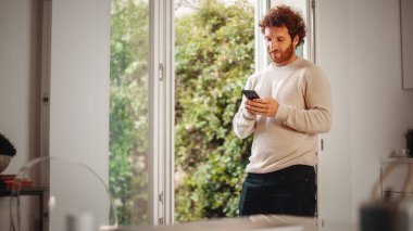 Ginger kıvırcık saçlı, akıllı telefon kullanan, Evdeki Oturma Odasında duran yakışıklı yetişkin adam. Erkek, bir fincan Espresso içip sosyal medyayı kontrol ediyor.