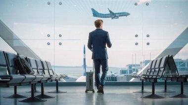 Havaalanı Terminali: Rolling Bavul Yürüyüşleri 'nde İşadamı, e-İş için Akıllı Telefon Uygulaması Kullanıyor. Gezgin Adam 'ın Geri Görüş Gelen Uçuşu Bekliyor