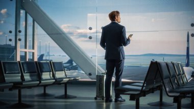 Havaalanı Terminali: Rolling Bavul Yürüyüşleri 'nde İşadamı, e-İş için Akıllı Telefon Uygulaması Kullanıyor. Geri Görünüm Gezgin Adam Uçuşu Bekliyor