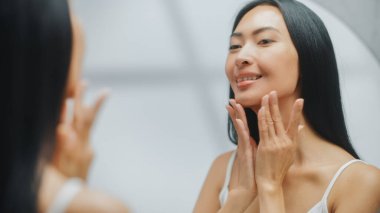 Güzel Asyalı Kadın 'ın portresi. Banyo aynasına nazikçe yüz kremi sürüyor. Genç Yetişkin Kadın Cildini Yumuşak, Yumuşak, Kırışıklıksızlaştırır