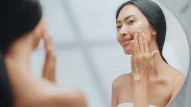 Güzel Asyalı Kadın Portresi. Nazikçe Yüz Kremi Maskesi ile Dokunarak Banyo Aynasına Bakıyor. Dişi, Tenini Yumuşatır