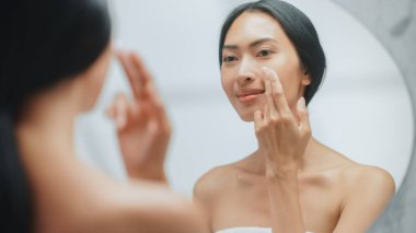 Güzel Asyalı Kadın Portresi. Nazikçe Yüz Kremi Maskesi ile Dokunarak Banyo Aynasına Bakıyor. Genç Dişi Cildini Yumuşattı