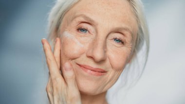 Güzel, kıdemli bir kadının portresi nazikçe yüz kremi sürüyor. Yaşlı kadın cildini yumuşatıyor, yumuşatıyor, kırışıklıkları yok ediyor ve doğal yaşlanmayı önlüyor.