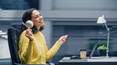 Modern Ofis: Gülümseyen İspanyol İş kadını Masasında oturuyor Bilgisayar üzerinde çalışıyor, kulaklık takıyor, Müzik dinliyor, dans ediyor. O mutlu, eğleniyor.