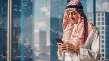 Geleneksel Beyaz Kandura 'da Genç Müslüman İşadamı, Modern Ofisinde, Gökdelenli Akıllı Telefon Kullanıyor. Başarılı.