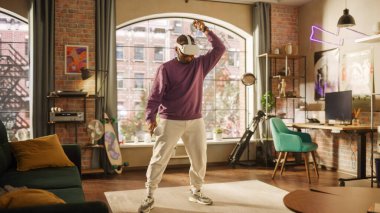 Genç erkek oyuncu evinde eğleniyor ve sanal gerçeklik oyunu oynuyor. Oturma odasında. Siyah adam modern bir kulaklık takıyor ve kontrol cihazlarını kullanıyor.