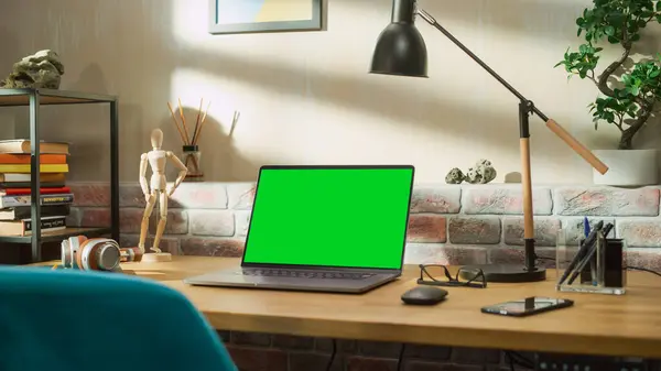 stock image Laptop Computer Standing on a Wooden Desk with a Green Screen Chromakey Mock Up Display. Cozy Empty Loft Apartment with a Lamp, Notebooks, Smartphone