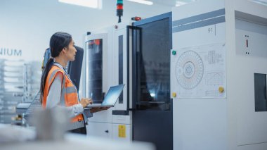Manufacturing Technician with a Laptop Comes Up to Operate a Heavy Industry Machine at a Factory. Asian Female Engineer Configuring Industrial