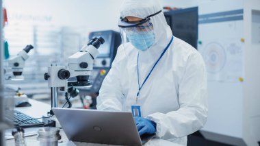 Electronic Manufacturing Factory: Engineer in Sterile Coverall Working on Laptop Computer, Examining a Circuit Board with Microchips Through a