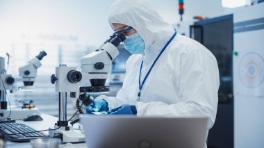 Electronic Manufacturing Factory: Engineer in Sterile Coverall Examining a Circuit Board with Microchips Through a Microscope. Specialist Comparing