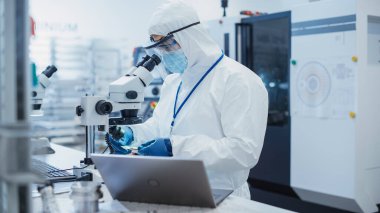 Portrait of an Engineer in White Safety Coverall Working on Laptop Computer at Electronic Manufacturing Factory. Technician Working on Daily Tasks and