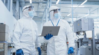 Two Scientists Walking in a Heavy Industry Factory in Sterile Coveralls and Face Masks, Using Laptop Computer. Examining Industrial Machine Settings