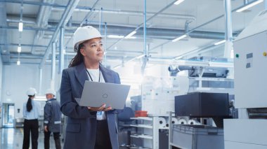 Portrait of a Black Female Engineer in Hard Hat Standing and Using Laptop Computer at Electronic Manufacturing Factory. Technician Thinking About