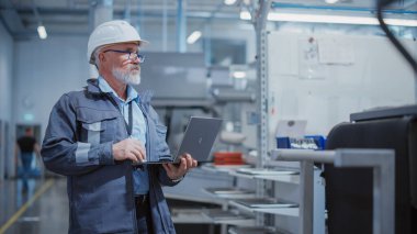 Bearded Middle Aged Chief Engineer Walking in a Factory Facility, Wearing a Work Suit, Stylish Blue Glasses and a White Hard Hat. Heavy Industry