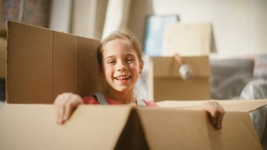 Portrait of Little Girl Plays Hide and Seek Jumping out of Cardboard Box, playing Hide and Seek. Cheerful Child Having Fun in Apartment Living Room
