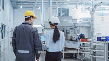 Back View: Two Heavy Industry Employees in Safety Uniforms and Hard Hats Walking and Talking in a Factory. Two Engineers Brainstorming and Solving