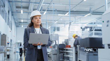 Portrait of a Happy Black Female Engineer in Hard Hat Standing and Using Laptop Computer at Electronic Manufacturing Factory. Technician Thinking
