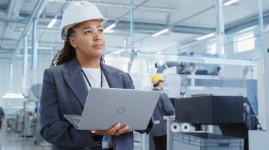 Portrait of a Black Female Engineer in Hard Hat Standing and Using Laptop Computer at Electronic Manufacturing Factory. Technician Thinking About