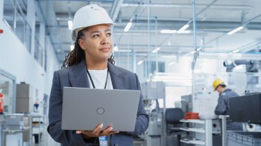Black African Female Engineer in Hard Hat Standing and Using Laptop Computer at Electronic Manufacturing Factory. Technician Working on Daily Tasks