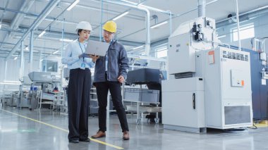 Two Young Asian Heavy Industry Engineers in Hard Hats Walking with Laptop Computer and Talking in a Factory. Two Manufacturing Employees at Work in