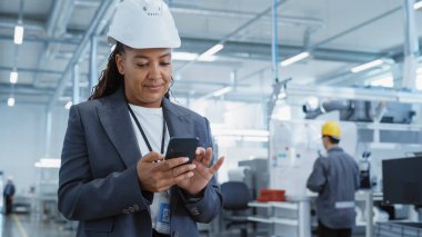 Portrait of a Black Female Engineer in Hard Hat Standing and Using a Smartphone at Electronics Manufacturing Factory. Technician is Writing a Message