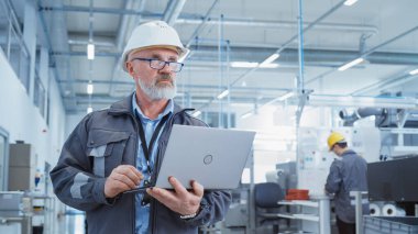 Portrait of a Bearded Middle Aged General Manager Standing in a Factory Facility, Wearing Work Jacket and a White Hard Hat. Heavy Industry Specialist