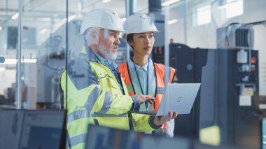 Two Professional Heavy Industry Employees Wearing Hard Hats at Factory. Checking and Discussing Work on Laptop Computer. Asian Quality Controller and