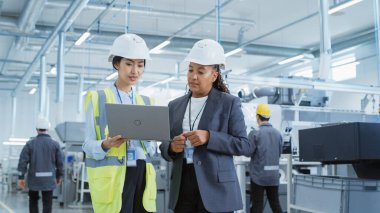 Two Female Heavy Industry Employees in Hard Hats at Factory. Discussing Job Assignments at Industrial Machine Facility, Using Laptop Computer. Asian