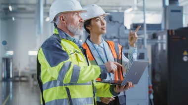 Two Diverse Professional Heavy Industry Engineers Wearing Safety Uniform and Hard Hats, Working on Laptop Computer. Asian Technician and Worker