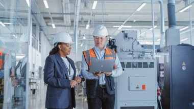 Two Professional Heavy Industry Engineers Walking at a Factory in Safety Uniform and Hard Hats, Discussing Work on Laptop Computer. Asian Specialist