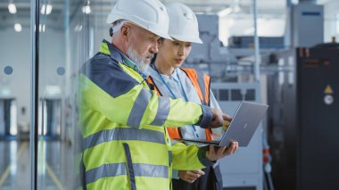 Two Diverse Heavy Industry Engineers in Safety Uniform and Hard Hats Walking with Laptop Computer and Talking in a Factory. Two Manufacturing