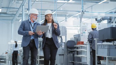 Two Heavy Industry Engineers in Hard Hats Walking with Laptop Computer and Talking in a Factory. Footage of Two Manufacturing Employees at Work in