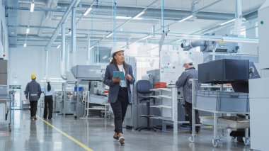 Portrait of a Black Female Engineer in Hard Hat Walking and Using Laptop Computer at Electronic Manufacturing Factory. Technician Working on Daily