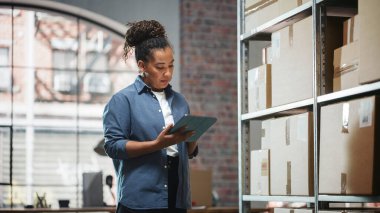 Inventory Manager Checks Stock, Writing in Clipboard Software on Tablet Computer. Black Woman Working in a Warehouse Storeroom with Rows of Shelves