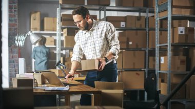 Inventory Manager Preparing a Small Cardboard Parcel for Postage. Stylish Young Male Small Business Owner Working on Laptop Computer in Warehouse