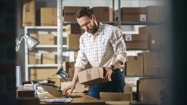 Small Business Owner of a Retail Online Shop Preparing a Small Cardboard Parcel for Postage. Stylish Young Inventory Manager Working on Laptop