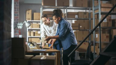 Multicultural Male and Female Warehouse Inventory Managers Talking, Using Laptop Computer and Checking Retail Stock. Rows of Shelves Full of Cardboard